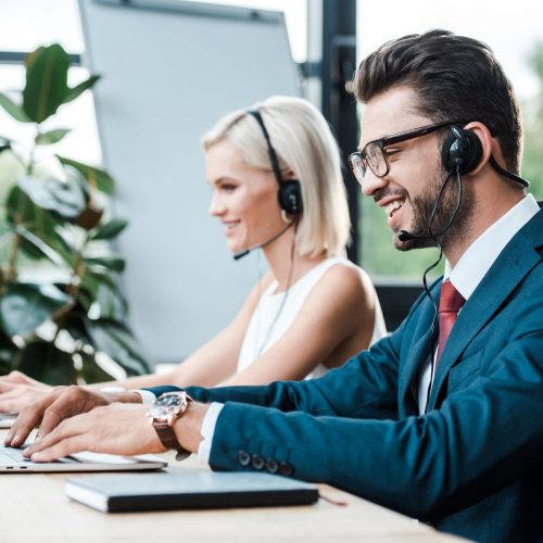 selective focus of happy operator in headset typing on laptop near blonde colleague in headset